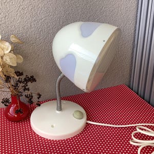 Peut inclure: Une lampe de table blanche avec un abat-jour en forme de champignon. L'abat-jour présente un motif en spirale bleu clair et blanc. La lampe a un col flexible et une base blanche avec un interrupteur rond.