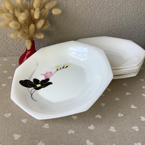 May include: White, octagonal plates with a floral design in black, pink, and yellow. A stack of plates is visible, suggesting a set. The plates are on a beige tablecloth with a heart pattern. A red vase with dried flowers is in the background.