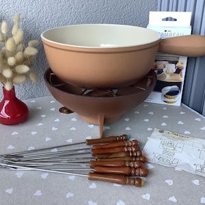 Può includere: Un set per fonduta vintage con una pentola e un supporto in ceramica marrone, un set di forchette per fonduta e un vaso rosso con fiori secchi. Il set include un foglio di istruzioni Le Creuset e una scatola di combustibile per fonduta.