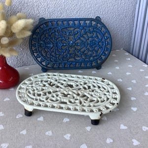 May include: Two decorative cast iron trivets. One is painted white with black feet, and the other is blue. Both have an oval shape with an ornate, openwork design. A red vase with dried flowers is in the background.