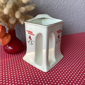 K&ouml;nnte beinhalten: Cremefarbener Keramikkrug mit dem "Johnnie Walker"-Logo in Rot. Der Krug hat eine quadratische Oberseite und ein griffartiges Design an den Seiten. Eine rote Vase mit getrockneten Blumen und orangefarbenen Kapseln ist ebenfalls zu sehen.
