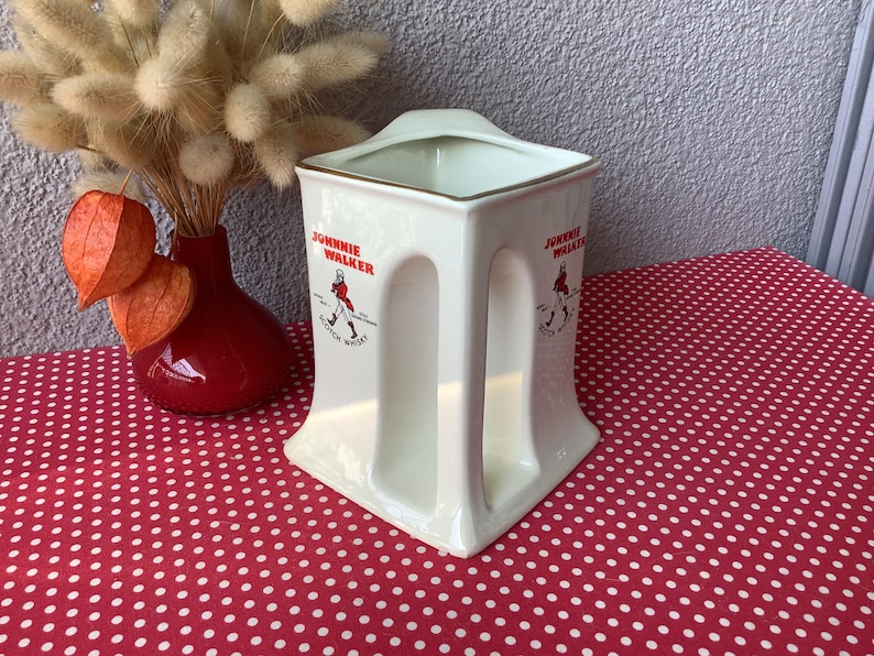 K&ouml;nnte beinhalten: Cremefarbener Keramikkrug mit dem "Johnnie Walker"-Logo in Rot. Der Krug hat eine quadratische Oberseite und ein griffartiges Design an den Seiten. Eine rote Vase mit getrockneten Blumen und orangefarbenen Kapseln ist ebenfalls zu sehen.