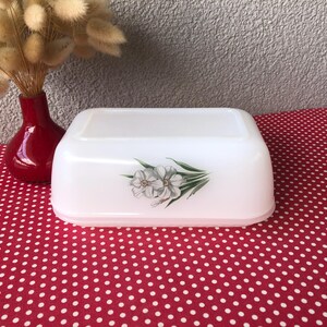 May include: A white rectangular butter dish with a floral design on the front. The dish is on a red polka dot tablecloth. A red vase with dried flowers is to the left of the dish.