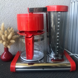 May include: A coffee maker with a bright red base, red filter housing, and a red handle on the glass carafe. The water reservoir is clear with black numerical markings from 1 to 12. The machine features a polished chrome finish on the base and top arm.