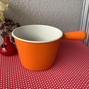 Può includere: Pentola per fonduta in smalto arancione con interno bianco e manico lungo. La pentola è appoggiata su una tovaglia rossa con pois bianchi.