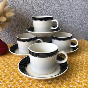 Op de afbeelding: Een set van vier witte keramische kopjes en schoteltjes met een zwarte rand en een klein wit ruitpatroon. Elk kopje heeft een oortje en een subtiel grijs verloop bij de basis. De schoteltjes hebben een bijpassende zwarte rand met lijnen.
