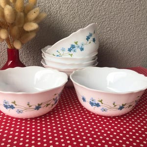 May include: A set of white glass bowls with scalloped edges, featuring a delicate floral pattern of small blue flowers and green stems wrapped around the exterior. The bowls are arranged in a stack and two individual bowls in the foreground.