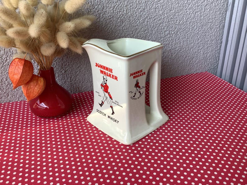 K&ouml;nnte beinhalten: Cremefarbener Keramikkrug mit dem "Johnnie Walker"-Logo und einer roten Figur. Der Krug hat einen quadratischen Sockel und einen Henkel. Der Text "Scotch Whisky" steht unter dem Logo. Eine rote Vase mit Trockenblumen befindet sich im Hintergrund.