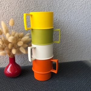 Puede incluir: Una pila de cuatro tazas de plástico en amarillo, verde oliva, blanco y naranja, cada una con un asa cuadrada. Junto a la pila hay un pequeño jarrón rojo que contiene un grupo de tallos de hierba de cola de conejo secos y esponjosos.