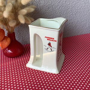 K&ouml;nnte beinhalten: Cremefarbener Keramikkrug mit dem Johnnie Walker-Logo und einer rot gekleideten Figur. Der Krug hat einen goldenen Rand und ein einzigartiges Griffdesign. Er steht auf einer roten Oberfl&auml;che mit wei&szlig;en Punkten, mit einer roten Vase und Trockenblumen im Hintergrund.