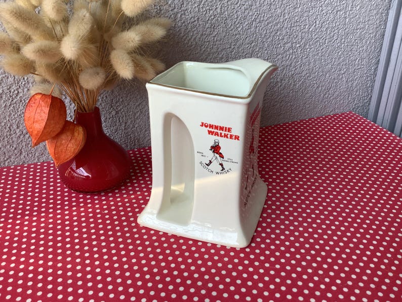 K&ouml;nnte beinhalten: Cremefarbener Keramikkrug mit dem Johnnie Walker-Logo und einer rot gekleideten Figur. Der Krug hat einen goldenen Rand und ein einzigartiges Griffdesign. Er steht auf einer roten Oberfl&auml;che mit wei&szlig;en Punkten, mit einer roten Vase und Trockenblumen im Hintergrund.