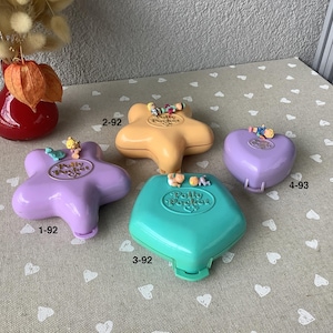 Può includere: Quattro compatti vintage di Polly Pocket in varie forme e colori, tra cui una stella, un cuore e un diamante. I compatti sono in tonalità di viola, giallo e verde acqua, con piccole figure sopra. L'immagine include anche un vaso rosso con fiori secchi.