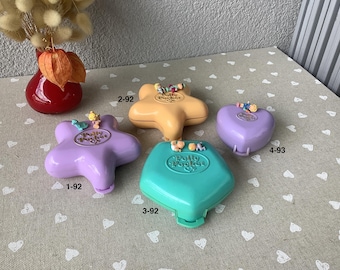 May include: Four vintage Polly Pocket compacts in various shapes and colors, including a star, heart, and diamond. The compacts are in shades of purple, yellow, and teal, with small figures on top. The image also includes a red vase with dried flowers.