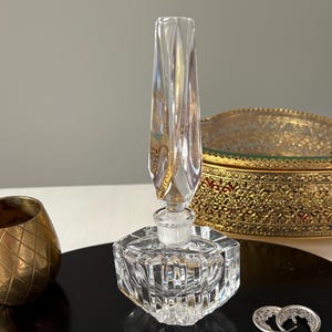May include: A clear crystal perfume bottle with a faceted base and a tall, decorative stopper. A gold-colored metal container and a pair of silver hoop earrings are also on display. The items are arranged on a black, round tray.