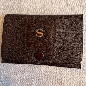 May include: A dark brown leather wallet with a textured surface. The wallet has a flap with a gold-colored "S" in a circular emblem. A small, round button secures the flap.