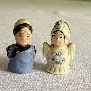 May include: Two small, hand-painted figurines of Asian figures. One figure wears a blue robe and has a black hair band. The other figure is in a cream-colored outfit with blue floral accents. Both have detailed facial features.