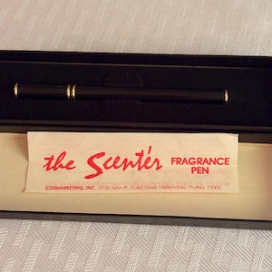 May include: A slim black fragrance pen with gold-tone accents, presented in a black box. A paper insert reads: the Scenter FRAGRANCE PEN, COSMARKETING, INC. 3136 John P. Curci Drive, Hallandale, Florida 33009. The pen has a sleek, cylindrical design.
