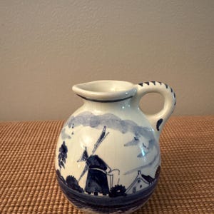 May include: A small, white ceramic pitcher with a blue windmill and landscape design. The pitcher has a rounded body, a handle, and a spout. The windmill is depicted with detailed blades and a small house. The handle is decorated with blue accents.