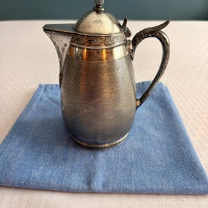 May include: An antique silver-plated coffee pot with a textured body, ornate handle, and a hinged lid. The pot is set on a small, square, light blue cloth napkin. The background is a light-colored tablecloth and a teal wall.