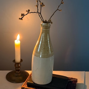 May include: A vintage stoneware bottle with a two-tone beige and white glaze, holding delicate branches with small buds. A lit candle in a brass candlestick and antique books complete the still life arrangement.