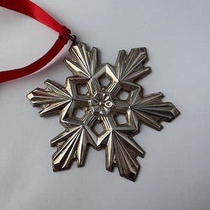May include: Silver-colored snowflake ornament with a red ribbon for hanging. The snowflake has a detailed, geometric design with six points. The ornament is likely made of metal and is suitable for holiday or winter decor.