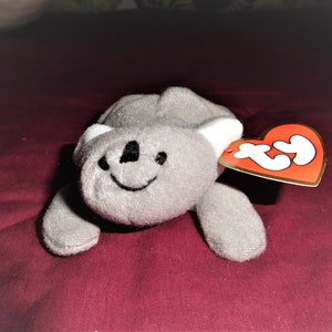 May include: A gray plush koala with a white belly and a black nose and mouth. The koala is lying on a red surface with a red Ty tag attached to its right arm.