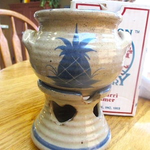 May include: A tan and blue ceramic oil warmer with a pineapple design. The warmer has a round top with handles and a base with heart-shaped cutouts. Blue stripes accent the base.
