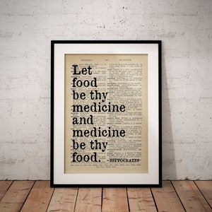 May include: A framed print displaying the quote "Let food be thy medicine and medicine be thy food" in black text. The quote is set against a vintage dictionary page background, with a black frame. The print is displayed on a wooden surface.
