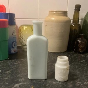 Könnte beinhalten: Eine hohe, rechteckige Flasche aus opakem weißem Glas mit engem Hals und ein kleineres, zylindrisches Gefäß aus opakem weißem Glas mit weiter Öffnung und schwach geprägter Schrift an der Seite, beide auf einer dunklen, gesprenkelten Oberfläche.
