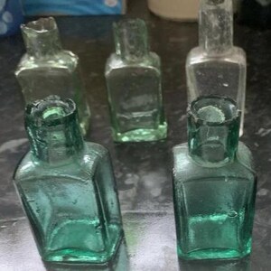 May include: A collection of five small, square-shaped glass bottles. Three of the bottles are a translucent light green colour, while two are clear glass. Each bottle features a short, narrow neck and a thick, rounded rim at the top opening.