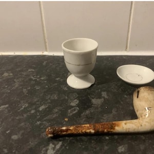 Może przedstawiać: Biały ceramiczny kieliszek do jajek z cienką złotą obwódką, mały biały ceramiczny spodek oraz zużyta fajka w kolorze złamanej bieli z brązowym, zardzewiałym ustnikiem. Fajka posiada ciemną, przetartą teksturę na całej długości ustnika.