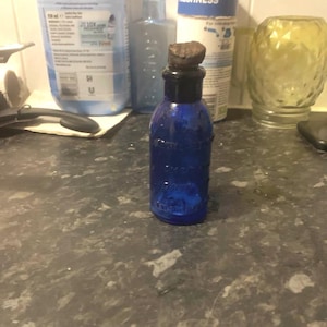 Könnte beinhalten: Eine kleine Apothekerflasche aus kobaltblauem Glas mit rundem Körper und kurzem, engem Hals. Die Flasche ist mit einem dunklen, strukturierten, zylindrischen Korkstopfen verschlossen. Auf der Vorderseite des Glases ist eine Prägung zu sehen.