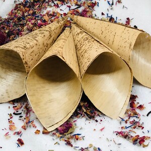 May include: Four natural brown paper cones, made from palm leaves, are arranged in a circle on a white surface. The cones are filled with dried flower petals in shades of pink, purple, and blue.
