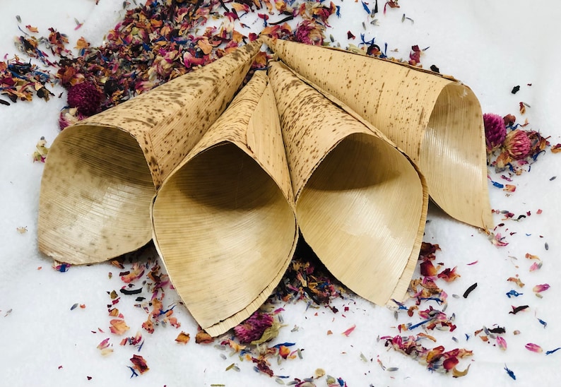 May include: Four natural brown paper cones, made from palm leaves, are arranged in a circle on a white surface. The cones are filled with dried flower petals in shades of pink, purple, and blue.