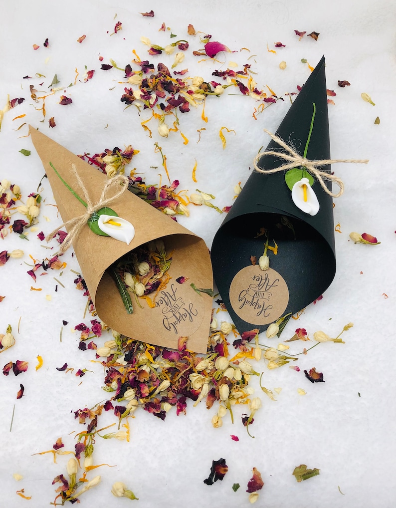 May include: Two brown paper cones filled with dried flower petals and a small white flower. The cones have a brown paper tag with the text "Happy Ever After" on them. The cones are surrounded by loose flower petals on a white background.
