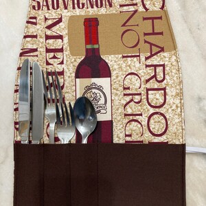 May include: A brown fabric utensil holder with a wine bottle design. The holder has four slots for silverware and the words "Chardonnay", "Pinot Grigio", "Merlot", and "Sauvignon" printed on the front.