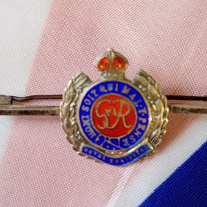 Peut inclure: Épingle argentée avec un emblème circulaire orné de détails en émail rouge et bleu. L'emblème est surmonté d'une couronne et entouré de texte. L'épingle est posée sur un fond rose, blanc et bleu.