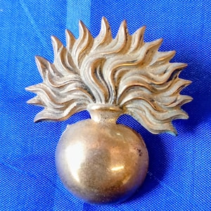 May include: A metallic gold-toned badge shaped like a spherical grenade with a stylized flame erupting from the top. The object features a smooth, rounded base and a textured, radiating flame design that fans out in a semi-circular pattern.