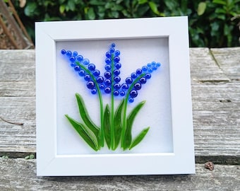 May include: A framed glass art piece featuring a blue flower design. The artwork showcases a cluster of blue glass beads forming the flower's blooms, with green glass leaves and stems. The white frame enhances the vibrant colors.