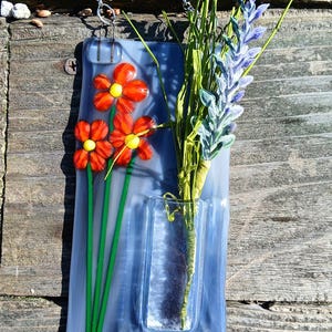 May include: A blue glass vase with a silver chain for hanging. The vase has a small opening for flowers and a painted design of red flowers with green stems. A bouquet of lavender and green foliage is in the vase.