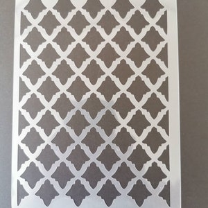 May include: A white plastic stencil featuring a repeating geometric pattern of interlocking squares and diamonds. The stencil is designed for crafting and decorating projects.