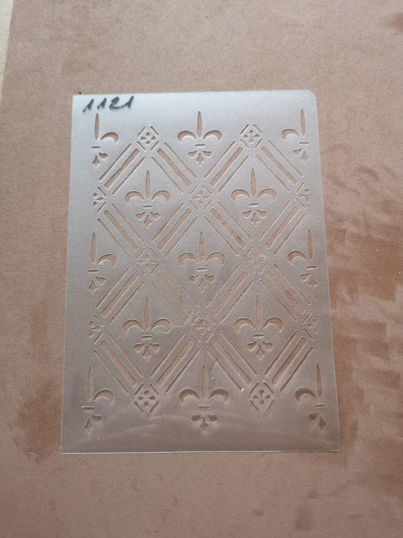 Peut inclure: Un pochoir en plastique transparent avec un motif r&eacute;p&eacute;titif de fleurs de lys et de formes de diamants. Le pochoir est &eacute;tiquet&eacute; "1121".