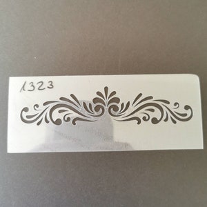 May include: A white plastic stencil with a decorative design. The stencil has the number "1323" written in the top left corner.