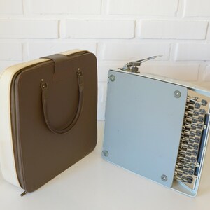 May include: A vintage brown and cream-colored typewriter case with a handle and a light blue typewriter. The case is rectangular with rounded corners. The typewriter is open, revealing the keys.