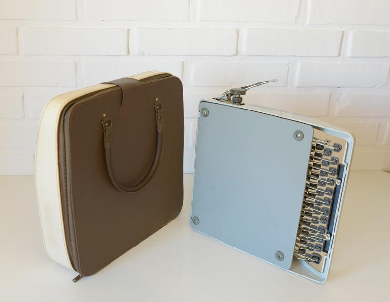May include: A vintage brown and cream-colored typewriter case with a handle and a light blue typewriter. The case is rectangular with rounded corners. The typewriter is open, revealing the keys.