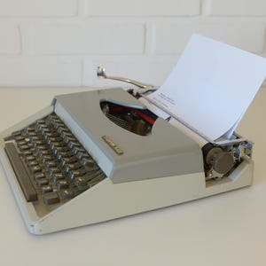 May include: A vintage gray and white typewriter with a blank white sheet of paper inserted. The typewriter has a full keyboard with black keys and a silver carriage return lever. The machine is on a white surface.