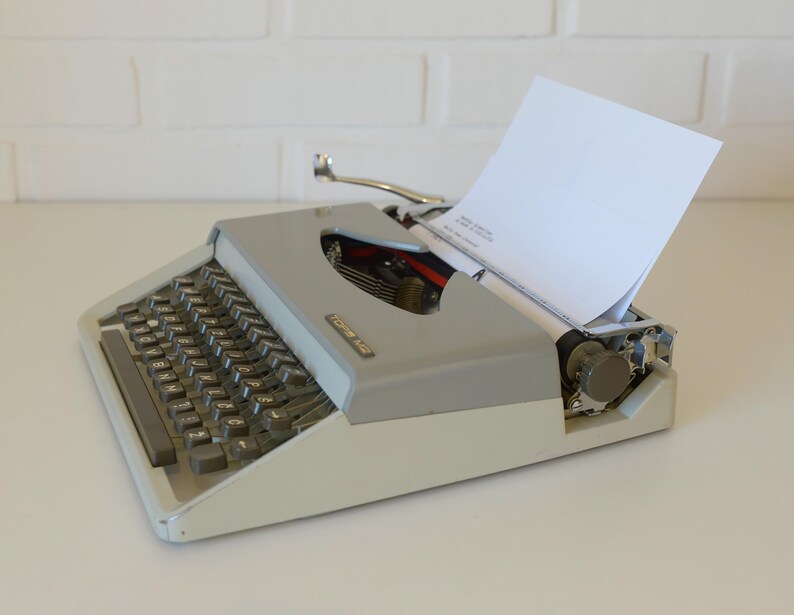 May include: A vintage gray and white typewriter with a blank white sheet of paper inserted. The typewriter has a full keyboard with black keys and a silver carriage return lever. The machine is on a white surface.