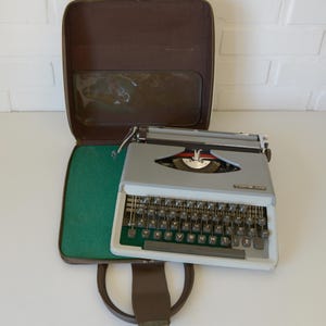 May include: A vintage gray typewriter with a white frame and black keys, resting on a green felt-lined case. The case is brown with a handle and a clear plastic window. The typewriter has a red and silver emblem.