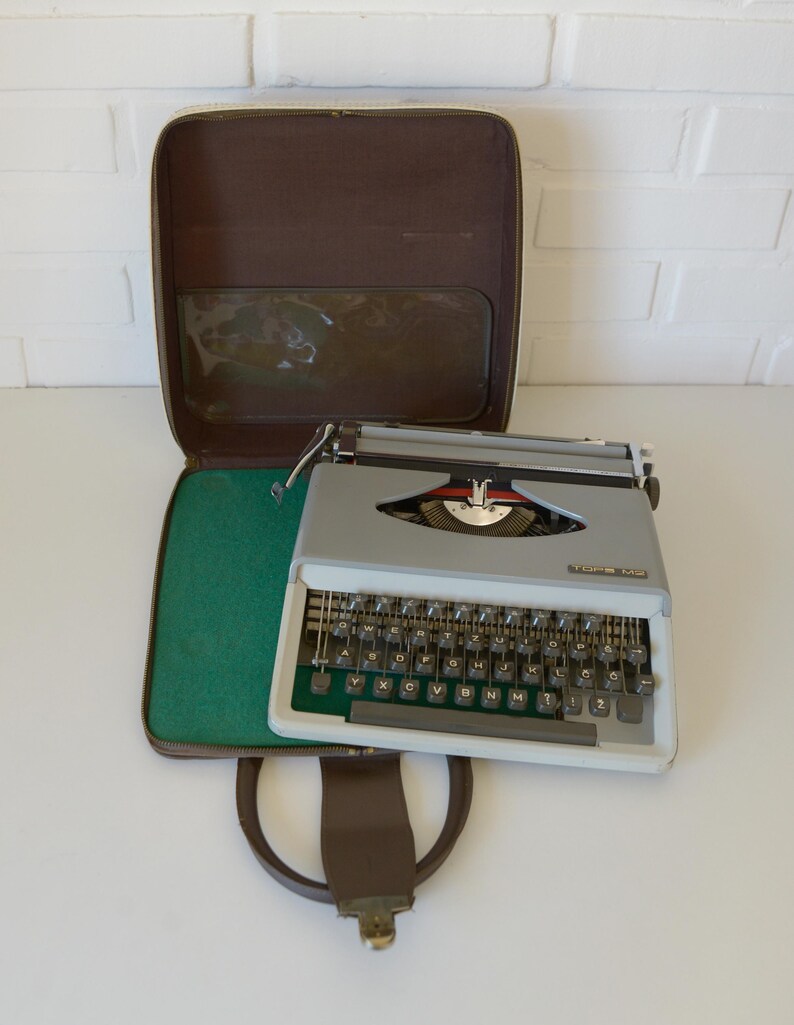May include: A vintage gray typewriter with a white frame and black keys, resting on a green felt-lined case. The case is brown with a handle and a clear plastic window. The typewriter has a red and silver emblem.