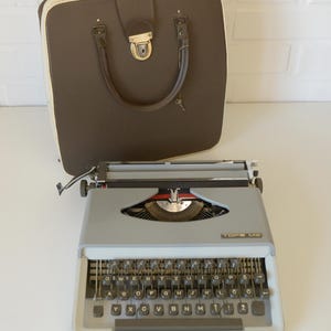 May include: A vintage gray TOPS M2 typewriter with a black keyboard and a brown carrying case. The typewriter has a silver carriage and a red ribbon indicator. The case is brown with a cream-colored trim and a handle.